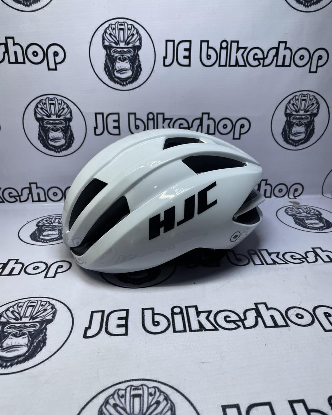 White bicycle helmet with black logo on a white background with 'JE bikeshop' branding.